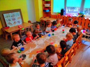 18 dzieci siedzi przy długim stole na którym rozsypana jest mąka.Dzieci bawiąc się mąka używają kolorowych kubków,miseczek,łyżek ,sitek .Z lewej strony stoi oparta o ścianę plansza obrazująca proces powstania pieczywa od zbioru zboża do gotowego produktu.