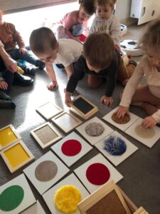 Grupka dzieci przygląda się tabliczkom z różnymi kolorowymi fakturami. Jeden chłopczyk pomaga rozkładać je na dywanie. Dziewczynka obiema rączkami sprawdza faktury na tabliczkach.